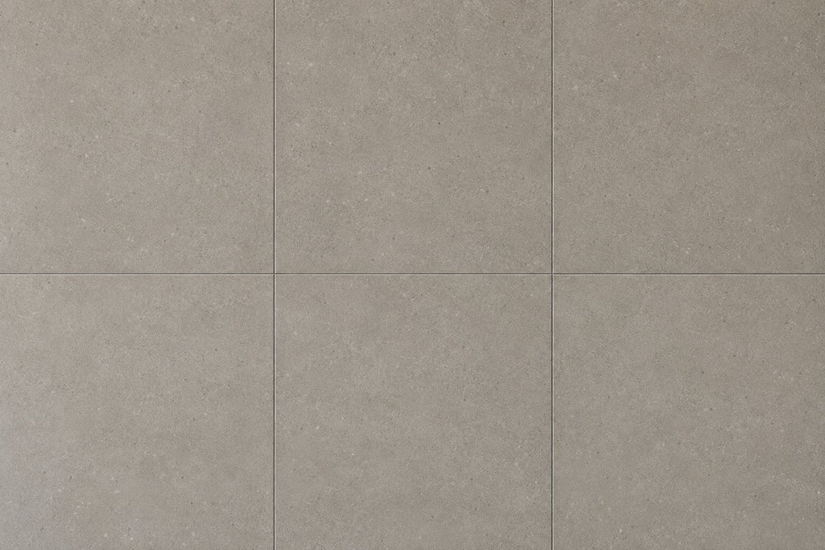 Editorial wall study: large square concrete-look tiles in a warm mid grey, laid in a grid with very fine joints, their flat powdery surface scattered with pale aggregate speckle under even daylight.