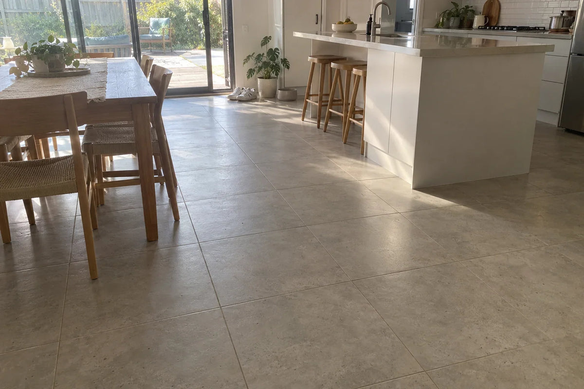 Open-plan kitchen and dining floor of large square pale greige concrete-look tiles with fine pale joints, an oak table, a white island bench and sun through a sliding door