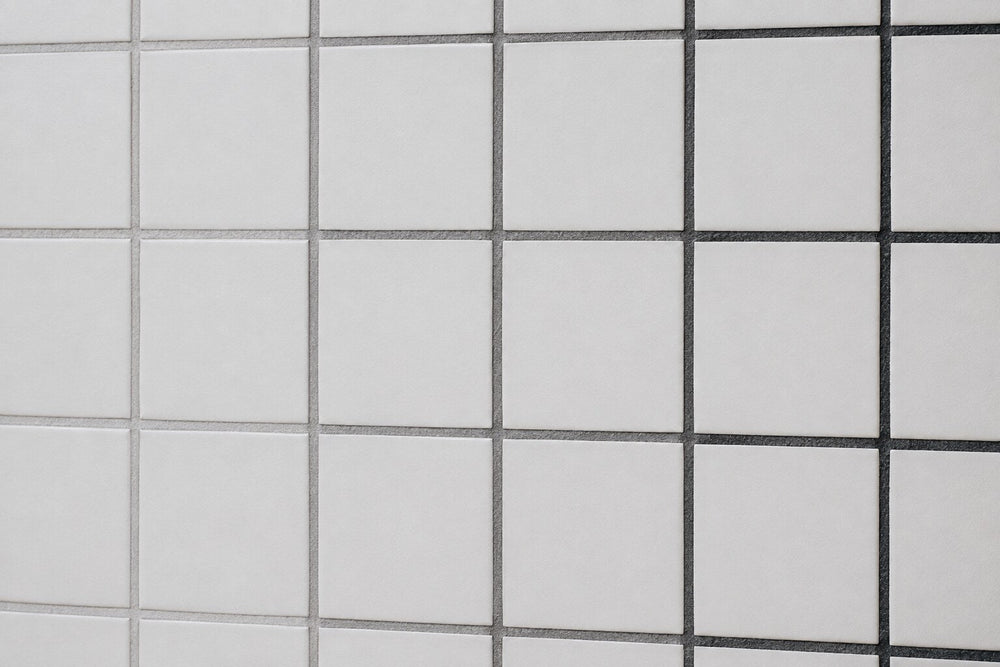 Editorial scene: plain white matt wall tiles with three adjacent runs of dried grout in light grey, mid grey and charcoal, showing how grout colour changes the same tile.