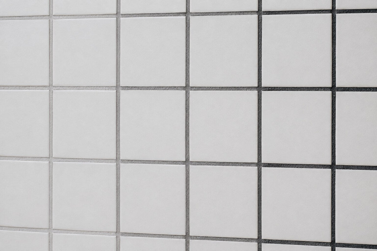 Editorial scene: plain white matt wall tiles with three adjacent runs of dried grout in light grey, mid grey and charcoal, showing how grout colour changes the same tile.