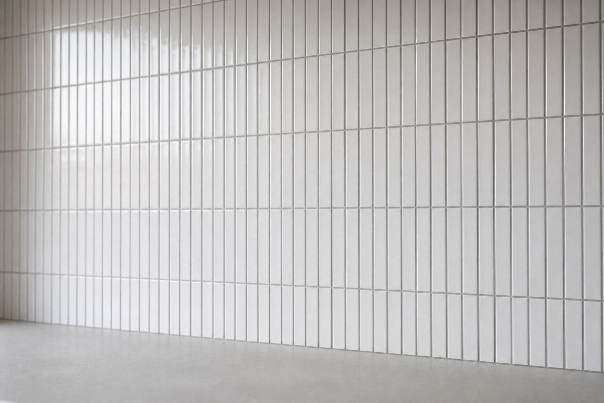 Editorial scene: slim white gloss finger mosaic tiles laid in a vertical stacked grid with grey grout above a plain bench, cool daylight.