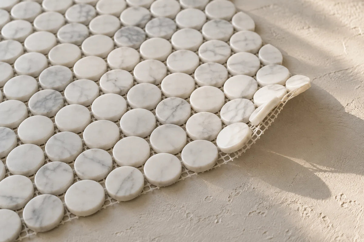 A sheet of small white marble penny round mosaic pieces on a mesh backing, lying on a pale plaster surface with one corner lifted so the mesh shows, the joints between the rounds still open.