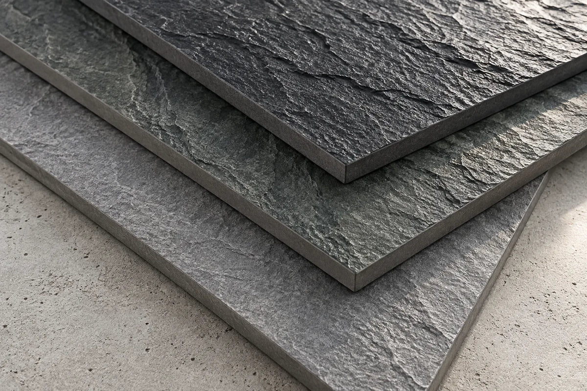 Corners of three slate-look tiles with a rough cleft face in charcoal, dark green-grey and mid grey, stacked on a concrete bench.