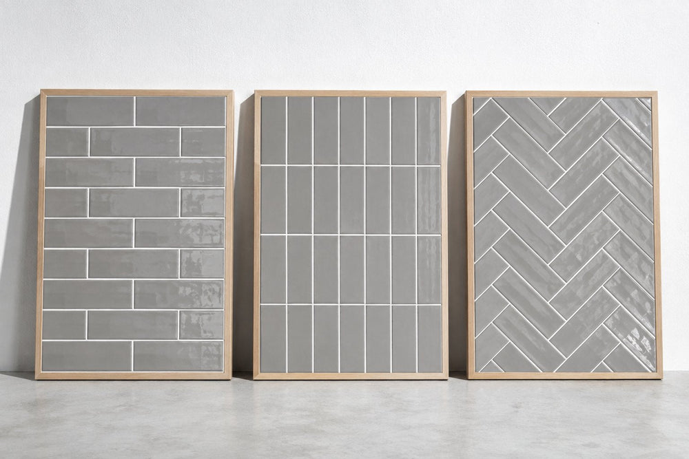 Three sample boards of grey gloss subway tile with white grout, laid in brick bond, vertical stack and herringbone, against a white wall in cool daylight.