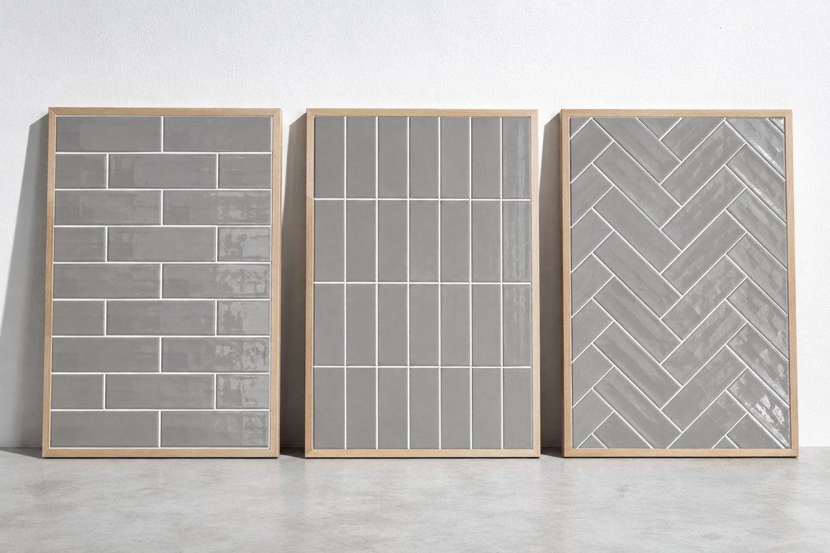 Three sample boards of grey gloss subway tile with white grout, laid in brick bond, vertical stack and herringbone, against a white wall in cool daylight.