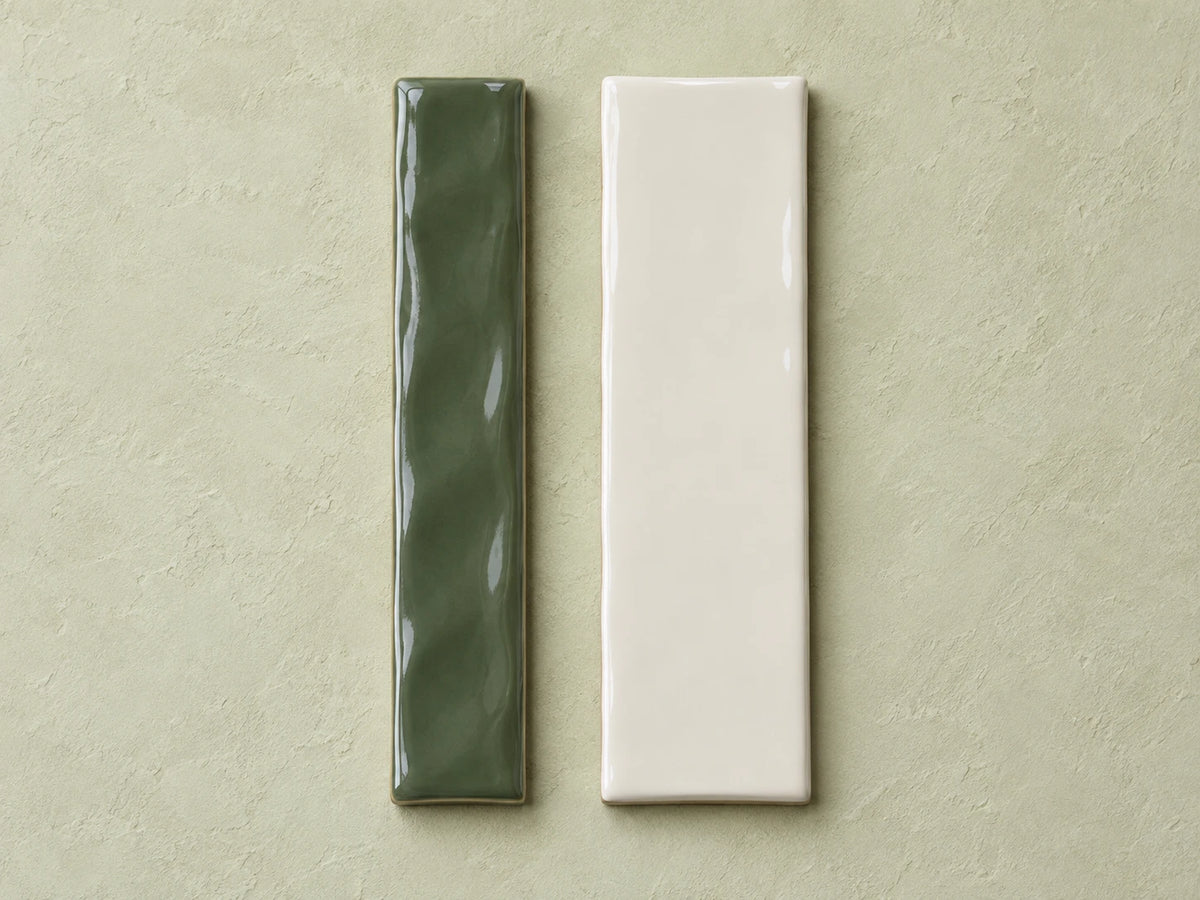Two gloss ceramic wall tiles on a pale sage plaster surface: a deep green one with an undulating wave face and a flat off-white one, the same length as each other but a different width.