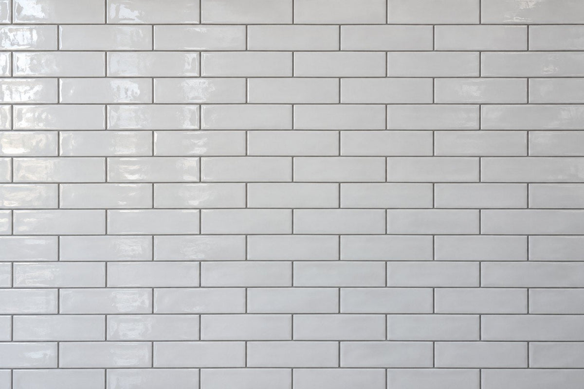 Editorial wall study: narrow white ceramic subway tiles laid in a running bond, their glossy uneven glaze catching daylight from the left, set with clearly visible mid-grey grout lines in a dense lattice.