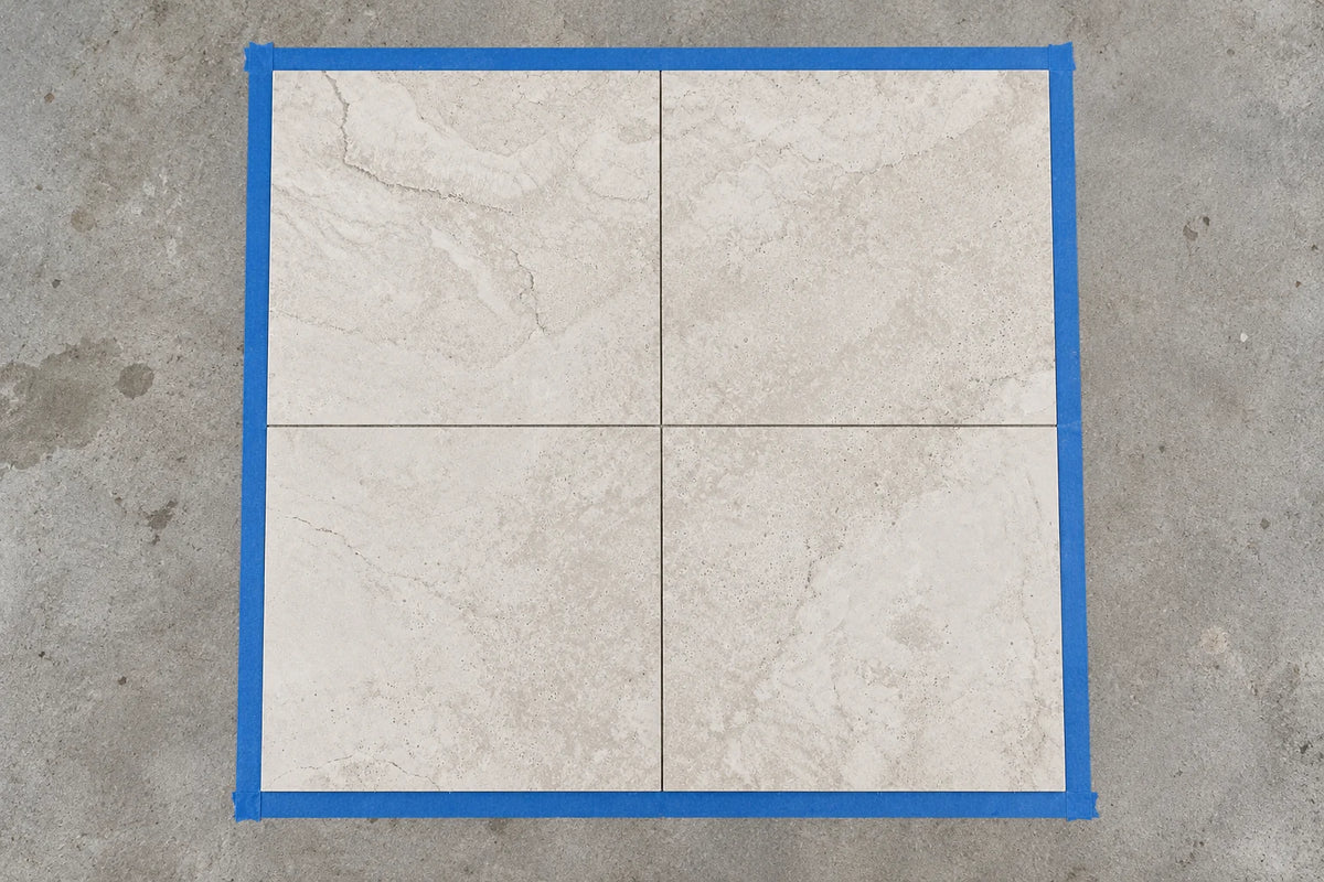 Four beige 600 × 600 mm tiles dry-laid inside a square of blue tape on a concrete slab