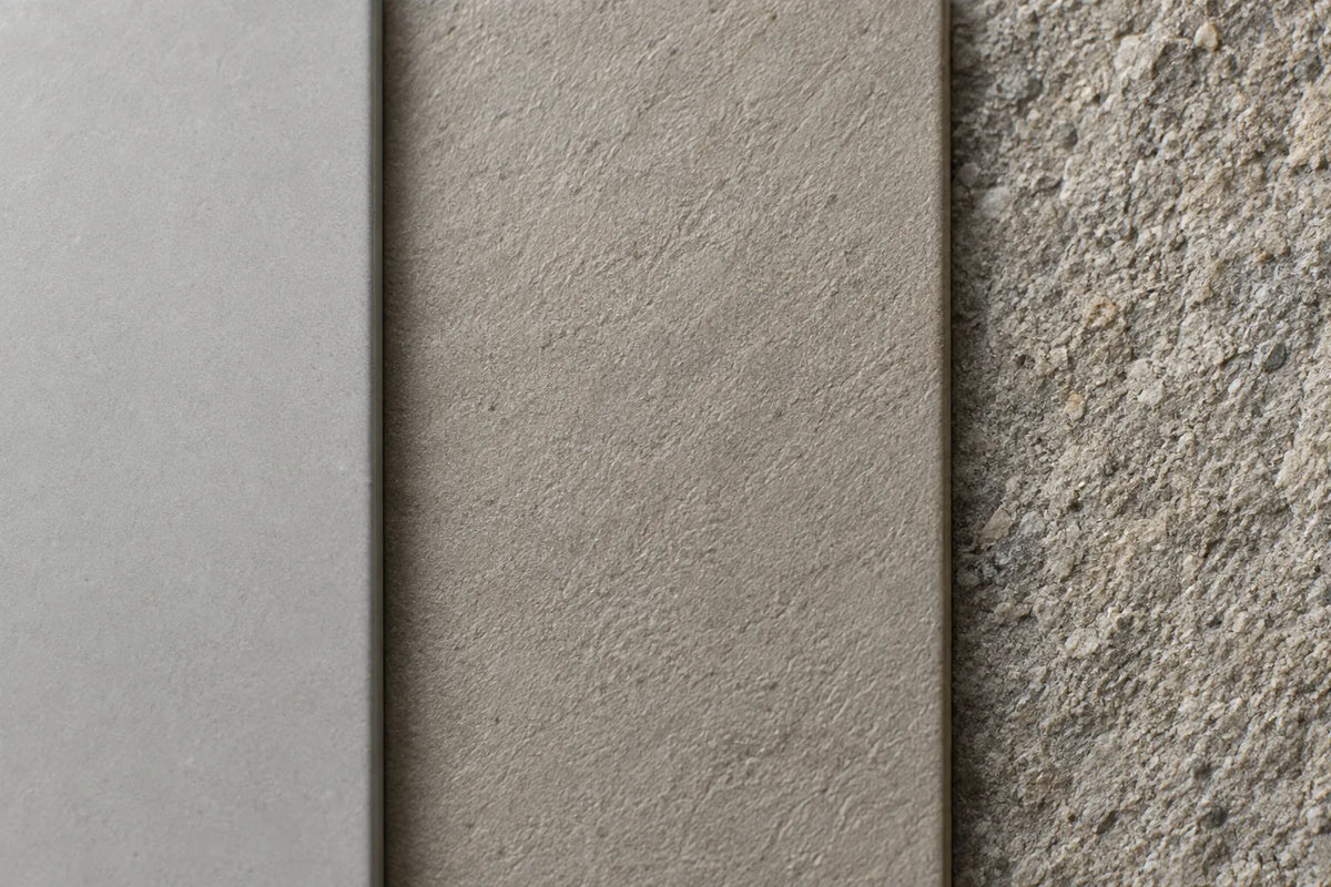 Close-up of three tile surfaces in low side light: smooth matt pale grey, lightly textured warm grey, and coarse and gritty.