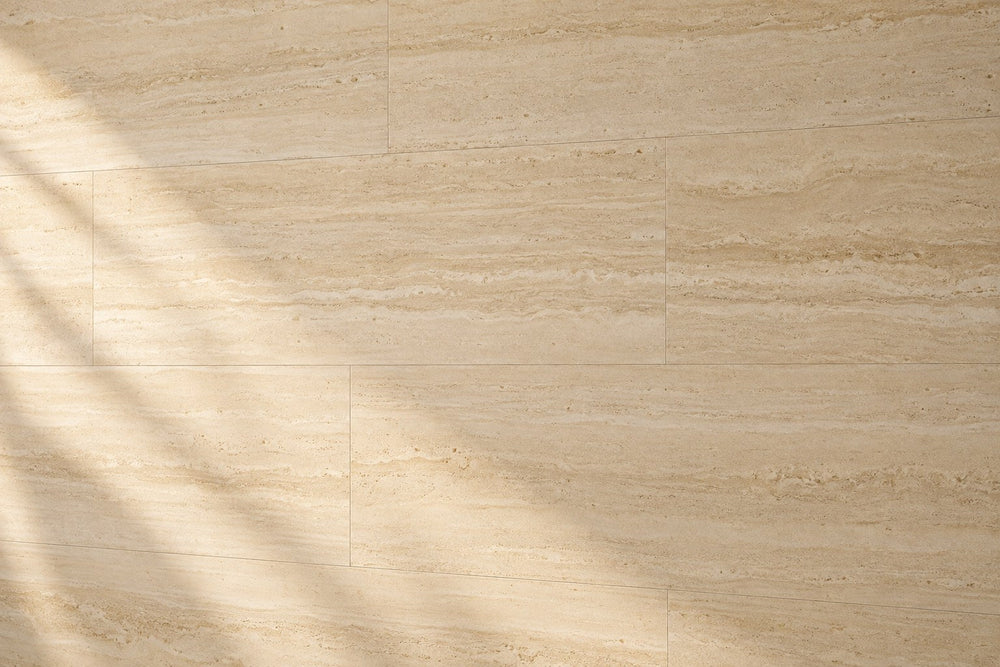 Editorial wall study: large ivory travertine-look tiles with a fine horizontal vein-cut grain, under soft late-afternoon daylight that throws a diagonal window shadow across the left. Quiet joints.