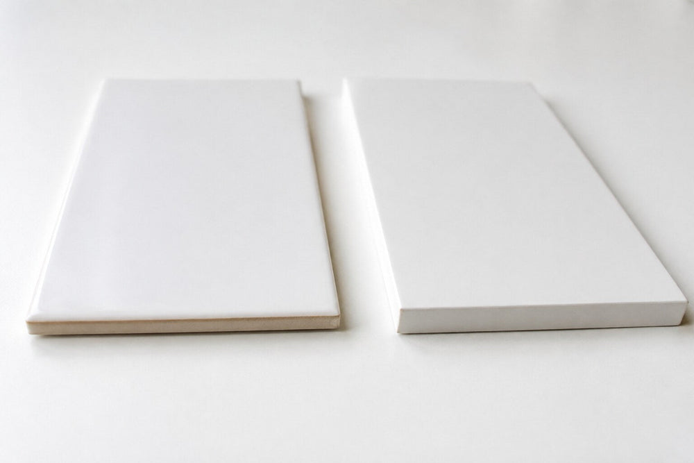 Editorial scene: two plain white tiles side by side on a white surface, one thinner with a glazed pillowed edge and one thicker with a sharp rectified edge.