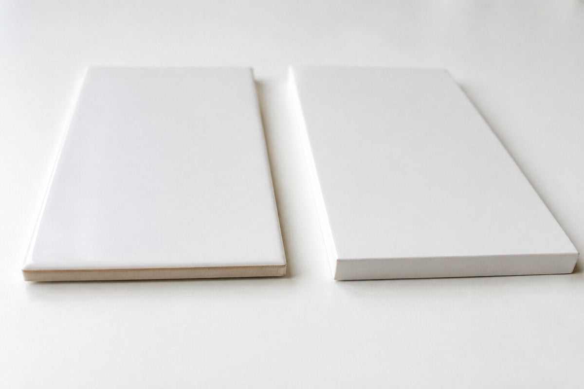 Editorial scene: two plain white tiles side by side on a white surface, one thinner with a glazed pillowed edge and one thicker with a sharp rectified edge.