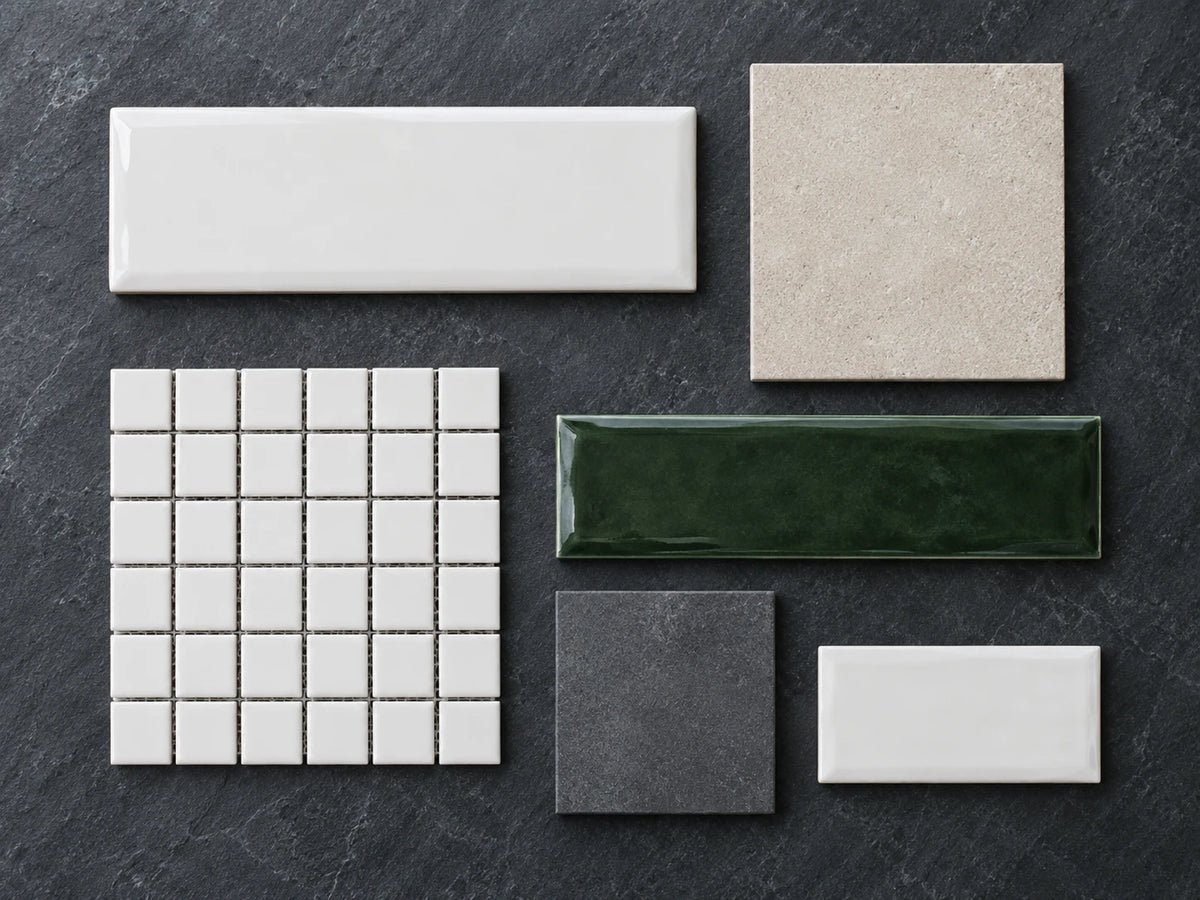 Six tile sample pieces of different sizes and finishes laid flat and separate on a dark charcoal slate bench, seen from overhead: gloss white and deep green rectangles, a beige square, a charcoal square and a white mosaic sheet.