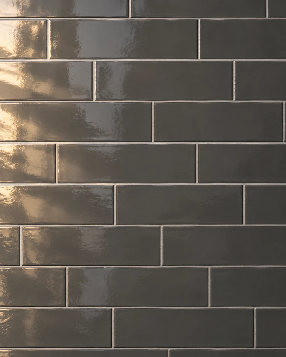 Charcoal gloss rectangular wall tiles laid so each course steps a third of a tile along from the one below, the joints forming a diagonal stair across the wall in warm raking light.