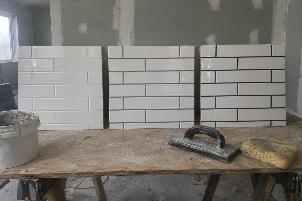 Three boards of white subway tiles on a work table in a renovation, grouted in white, mid grey and charcoal, with a grout float and sponge