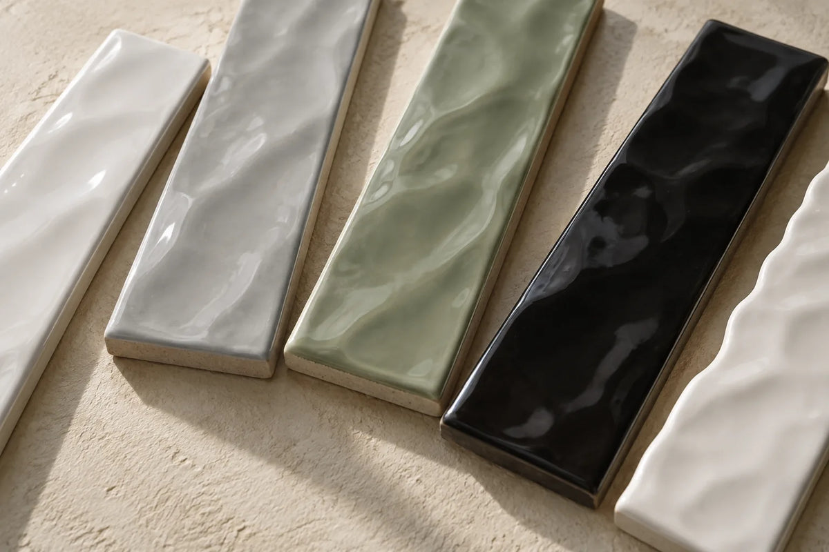 Five glossy wave-face subway tiles in white, grey, sage green, black and a ripple-edged white, set side by side on pale plaster in side light