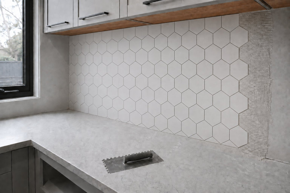 Kitchen splashback mid-installation: white matt hexagon tiles of about 100 mm over most of the wall, a narrow strip of combed grey adhesive still bare, joints ungrouted, a trowel on the bare bench. Generated illustration, not a Tilekind product.