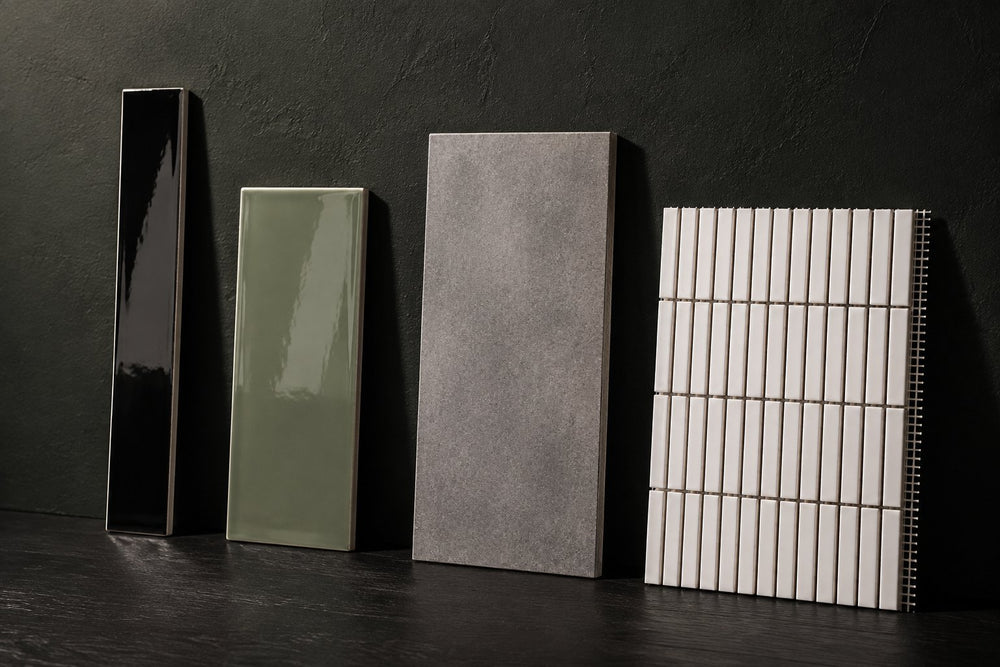 Four unbranded tile samples against a dark plaster wall: a narrow black gloss subway tile, a wider sage-green gloss subway tile, a large grey concrete-look tile and a white strip-mosaic sheet. Generated illustration, not Tilekind products.