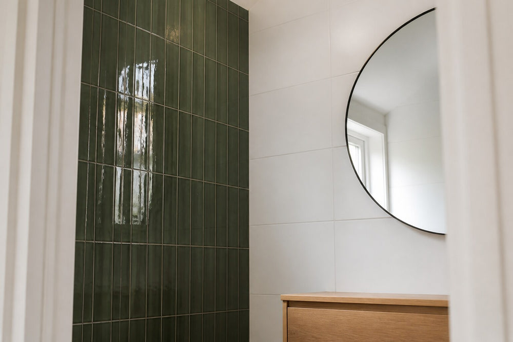 Editorial scene: a compact ensuite seen from the doorway, deep green gloss tiles stacked vertically on one wall, plain white tiles on the others, a timber vanity and round mirror. Generated illustration, not a Tilekind product.