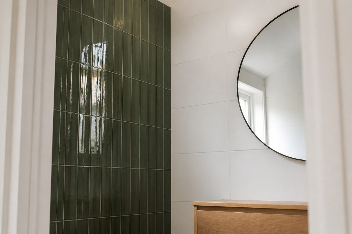 Editorial scene: a compact ensuite seen from the doorway, deep green gloss tiles stacked vertically on one wall, plain white tiles on the others, a timber vanity and round mirror. Generated illustration, not a Tilekind product.