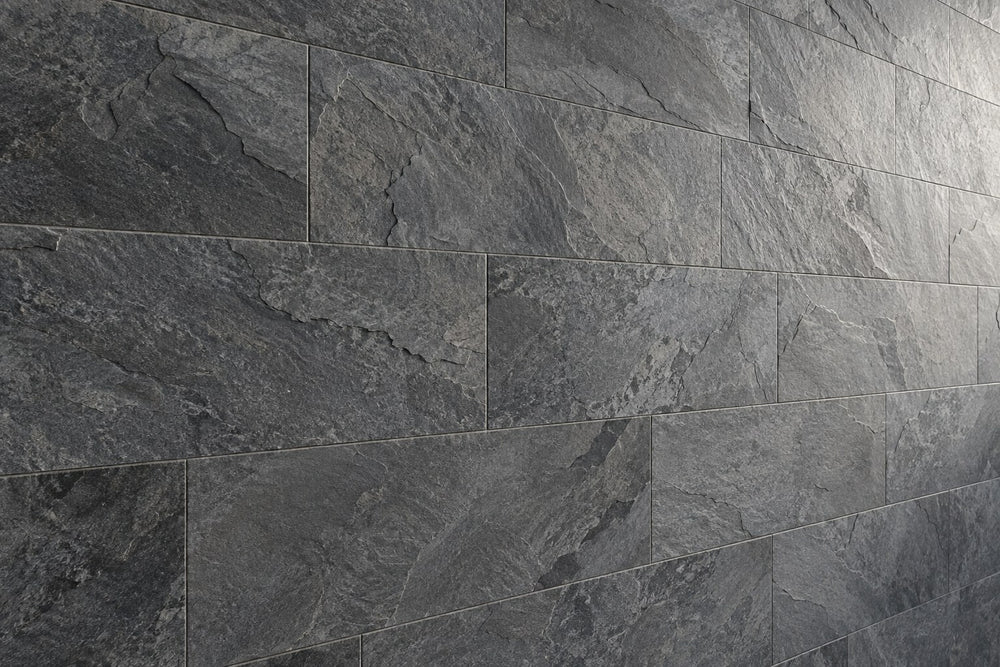 Editorial wall study: large rectangular stone-look tiles in charcoal and mid grey, laid in a running bond, their broken cleft surface picked out by raking daylight from the right. Narrow joints. Generated illustration, not a Tilekind product.