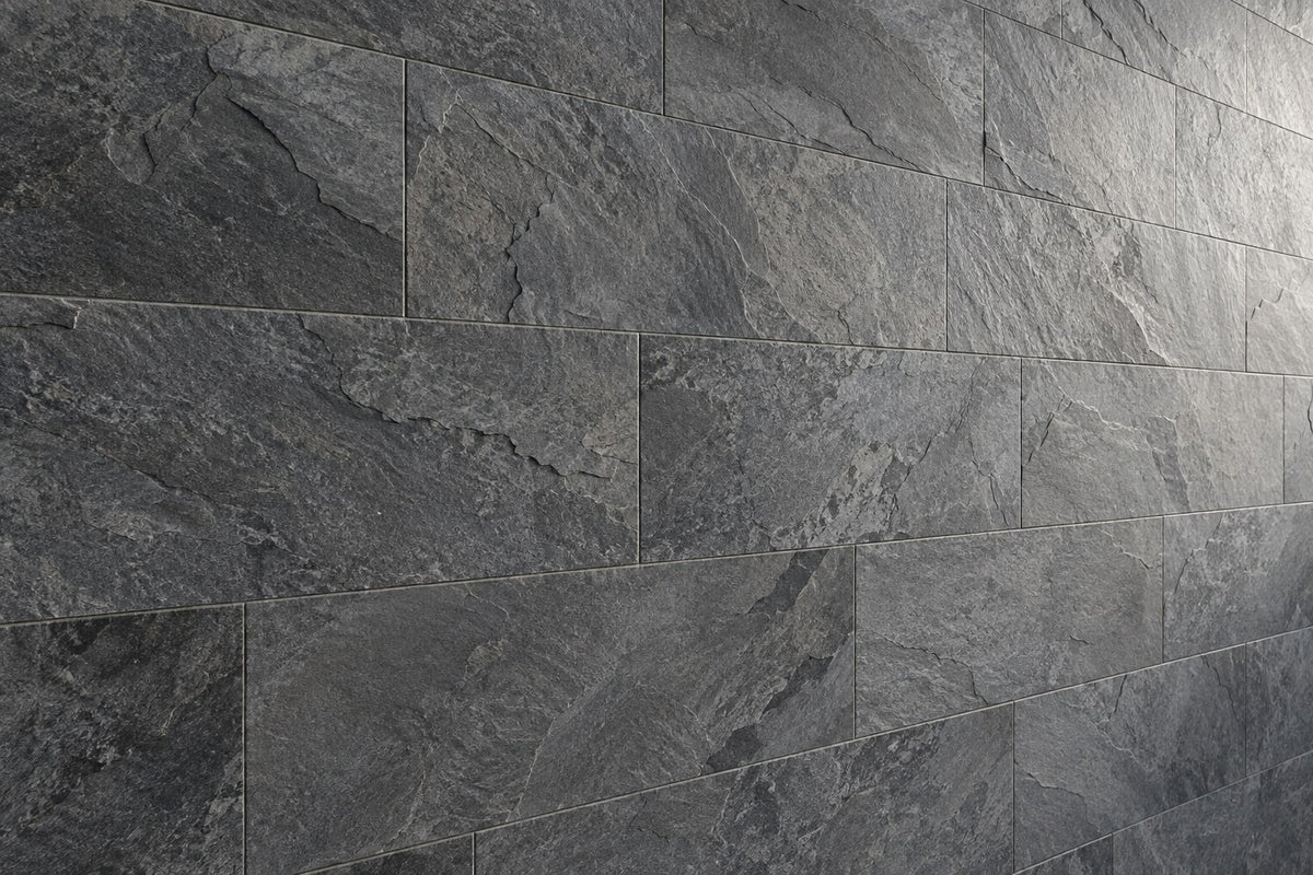 Editorial wall study: large rectangular stone-look tiles in charcoal and mid grey, laid in a running bond, their broken cleft surface picked out by raking daylight from the right. Narrow joints. Generated illustration, not a Tilekind product.