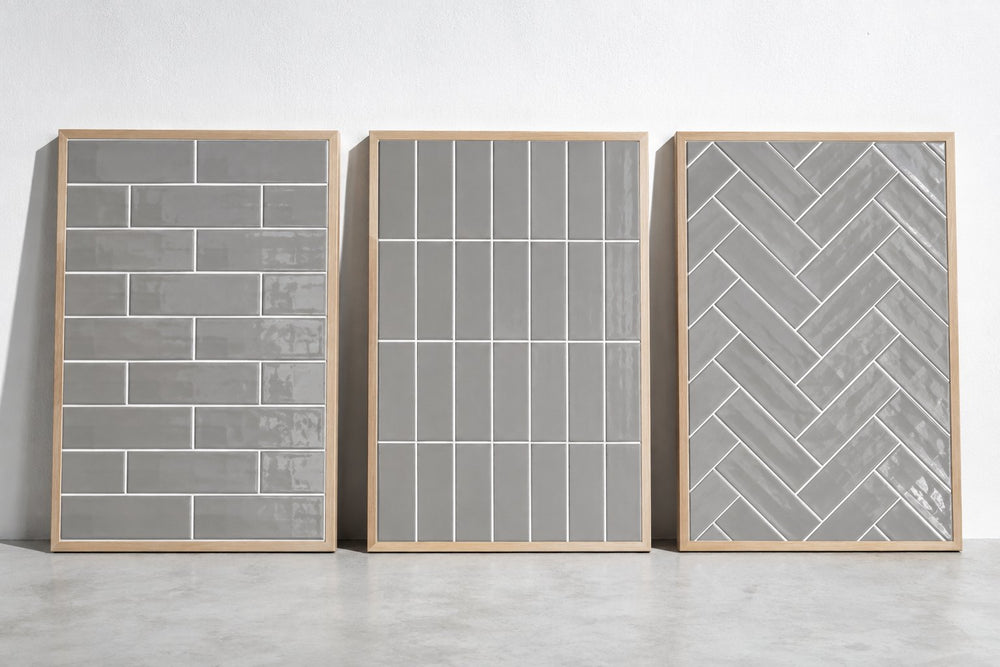Three sample boards of grey gloss subway tile with white grout, laid in brick bond, vertical stack and herringbone, against a white wall in cool daylight. Generated illustration, not a Tilekind product.