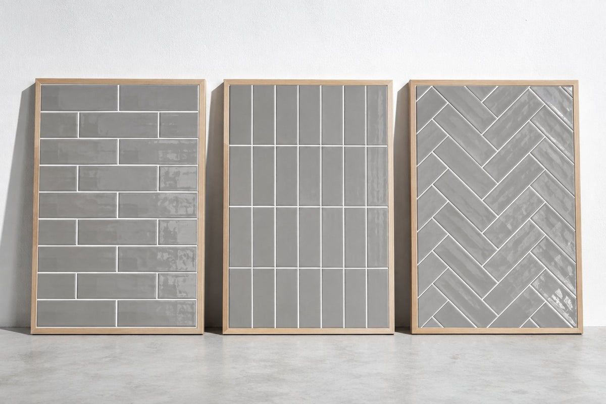 Three sample boards of grey gloss subway tile with white grout, laid in brick bond, vertical stack and herringbone, against a white wall in cool daylight. Generated illustration, not a Tilekind product.