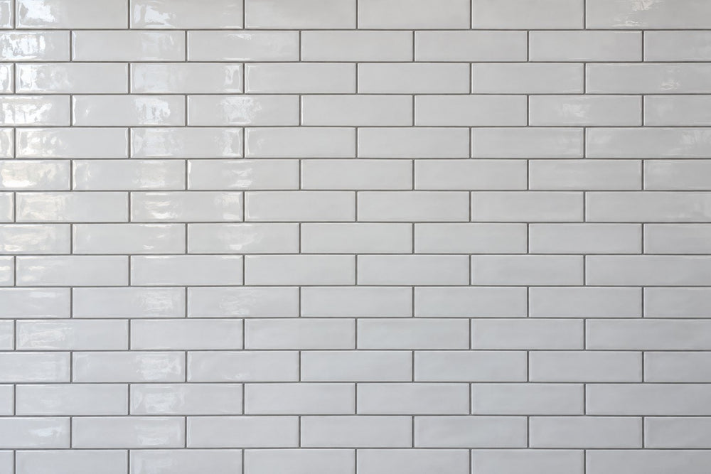 Editorial wall study: narrow white ceramic subway tiles laid in a running bond, their glossy uneven glaze catching daylight from the left, set with clearly visible mid-grey grout lines in a dense lattice. Generated illustration, not a Tilekind product.