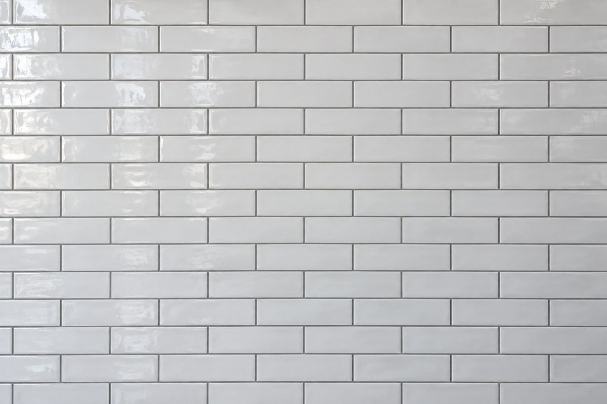Editorial wall study: narrow white ceramic subway tiles laid in a running bond, their glossy uneven glaze catching daylight from the left, set with clearly visible mid-grey grout lines in a dense lattice. Generated illustration, not a Tilekind product.
