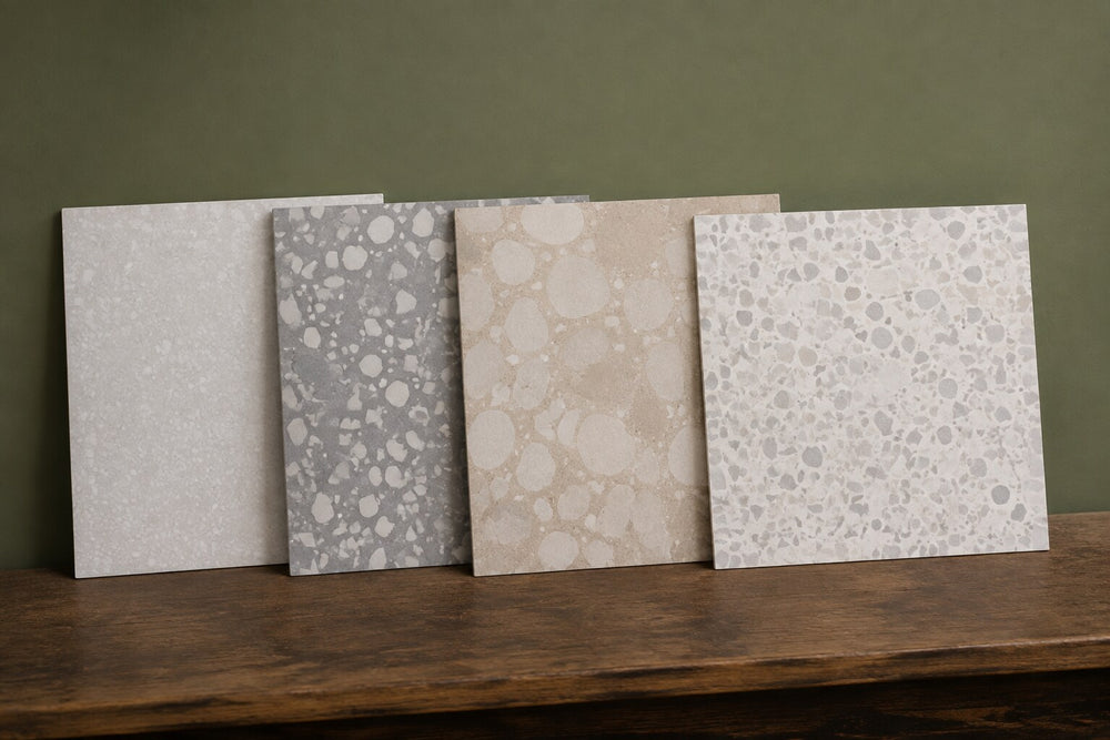 Editorial scene: four square terrazzo-look tiles with fine, large and oversized chips leaning against an olive wall on a dark timber bench. Generated illustration, not a Tilekind product.