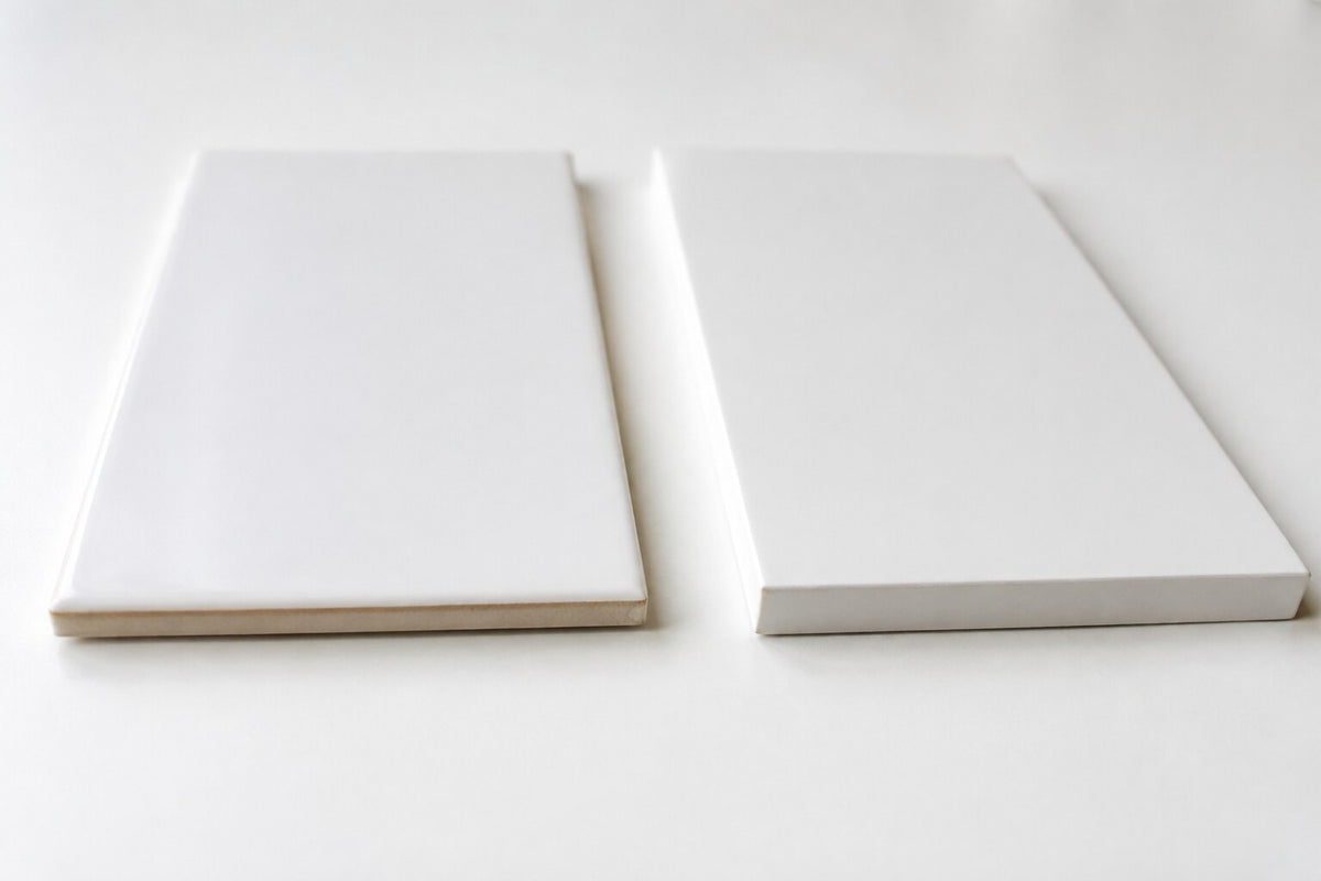 Editorial scene: two plain white tiles side by side on a white surface, one thinner with a glazed pillowed edge and one thicker with a sharp rectified edge. Generated illustration, not a Tilekind product.