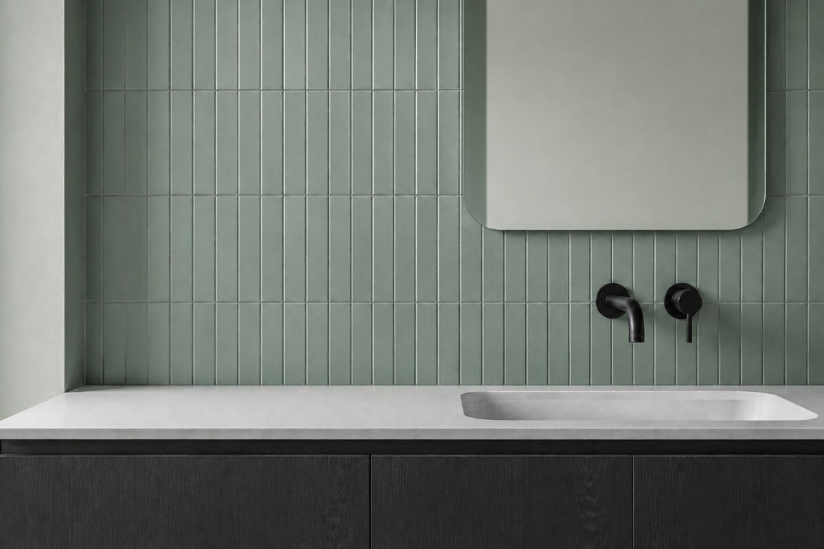 Muted sage-green vertical wall tiles sit behind a dark timber vanity with a pale basin, rounded mirror and matt-black wall tapware.