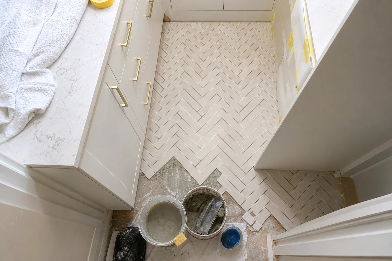 High-angle view of a pale herringbone mosaic floor being installed in a narrow kitchen